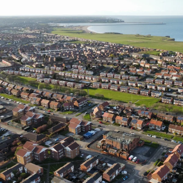 An aerial view shows a large residential area of houses with red rooftops, with a large green field separating the housing from the coastline. The sea is visible in the distance.