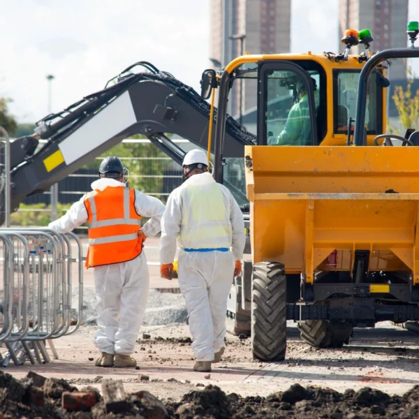 Two site workers in protective white coveralls and hard hats standing next to a yellow truck and an excavator at a construction site.